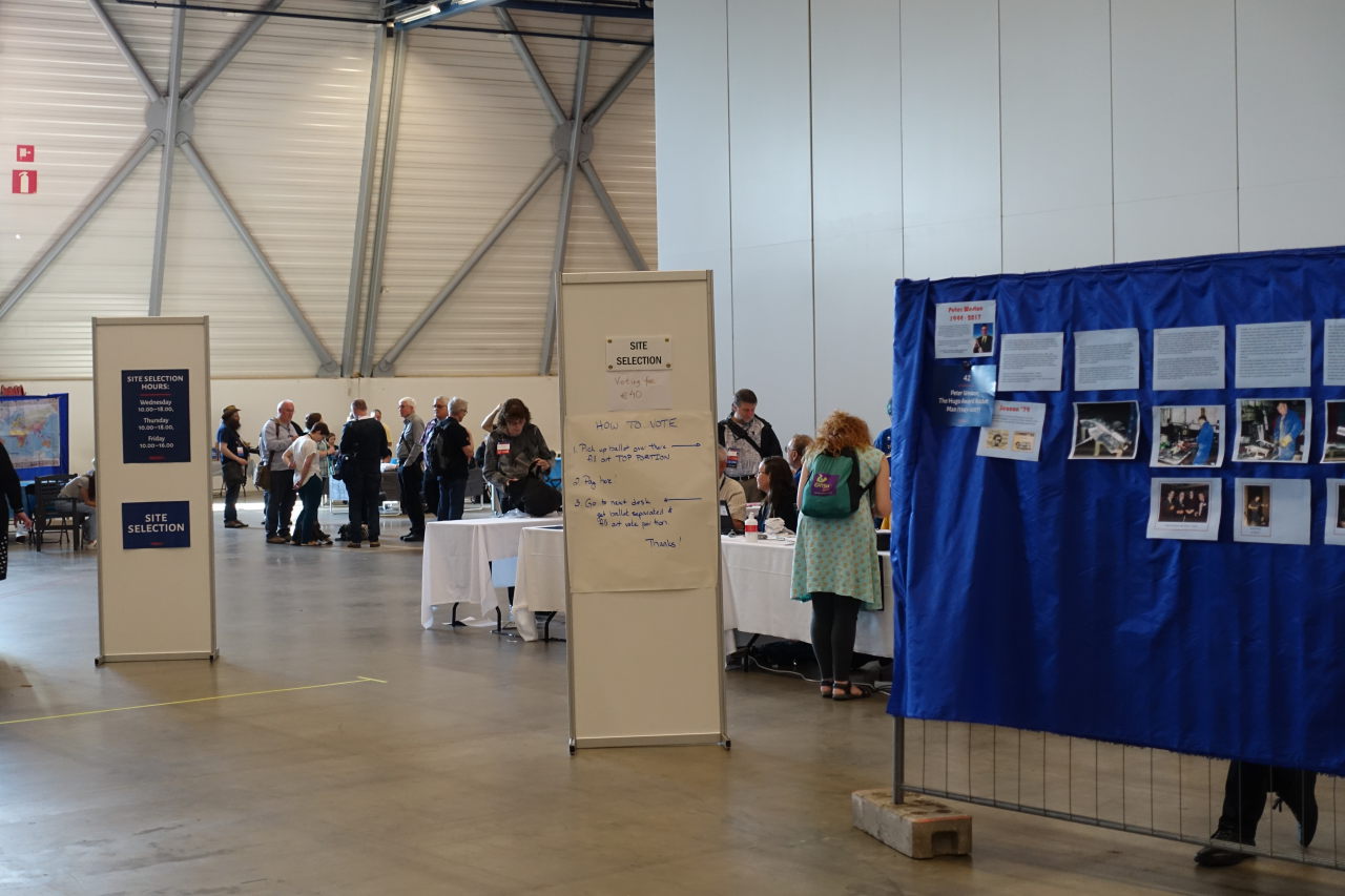 Interior of a big hall. To the right there is an exposition on dark blue fabric. In the center there are two stands with 'Site Selection' posters and information. Behind the stands there are people and tables.
