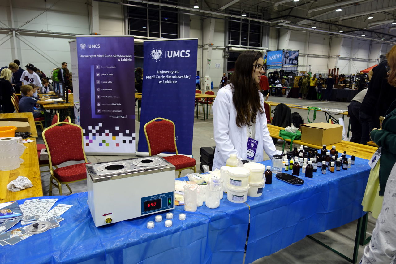 Woman in white lab coat standing behind the table full of chemical reagents in small bottles and plastic boxes. There is also water bath on the table.