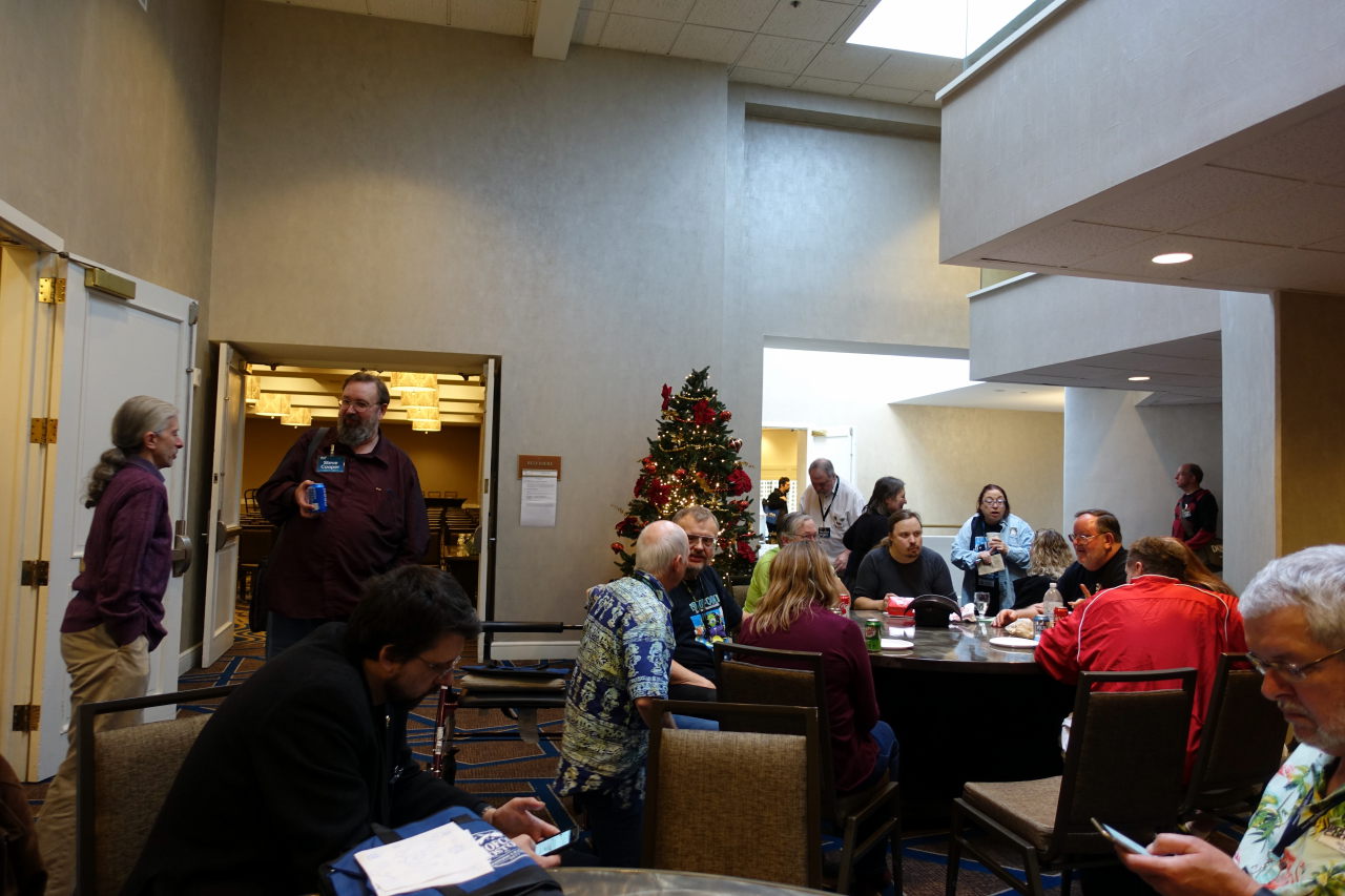 People are sitting around the round tables. Two man are standing near the door and talking. In the background christmas tree is visible.