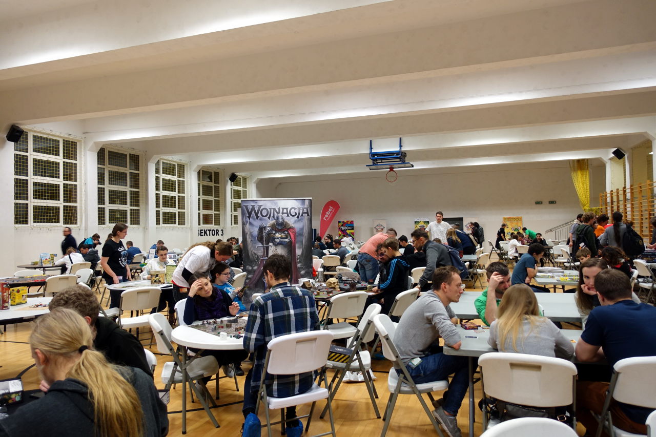 Big hall with multiple tables. People are sitting around the tables and are playing games.