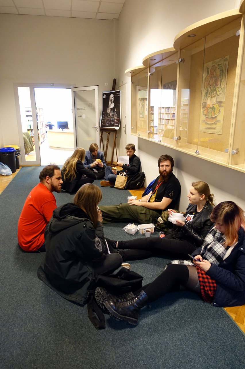 Eight people sitting on a floor in the corridor. A few of them are eating.