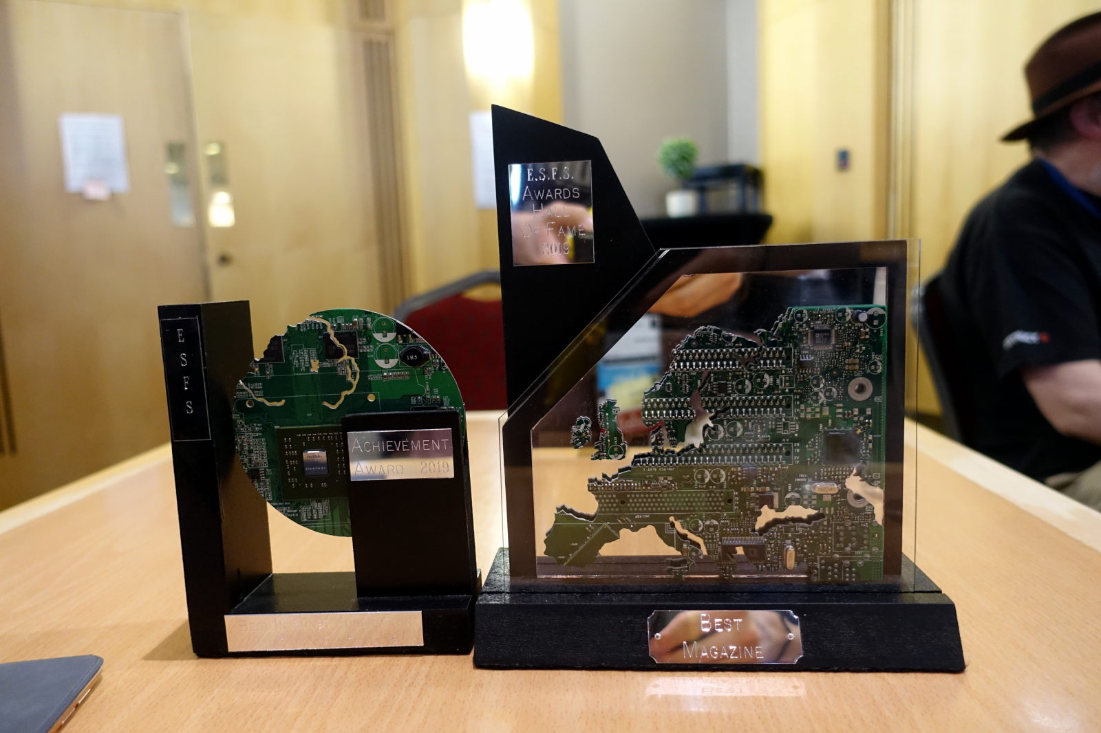 Two awards standing next to each other. To the lefy ESFS Achievement Award - made of round piece of circuit board in a wooden holder. To the right ESFS Award for Best Magazine - map of Europe cut out of circuit board on a wooden holder.