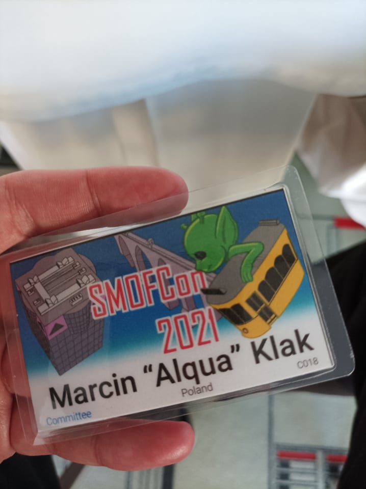 Convention badge hold in hand. The badge shows convention name ('SMOFCon 2021') and the name of the member. Picture on the badge depicts a tall modern building, aqueduct and an alien holding yellow tram.