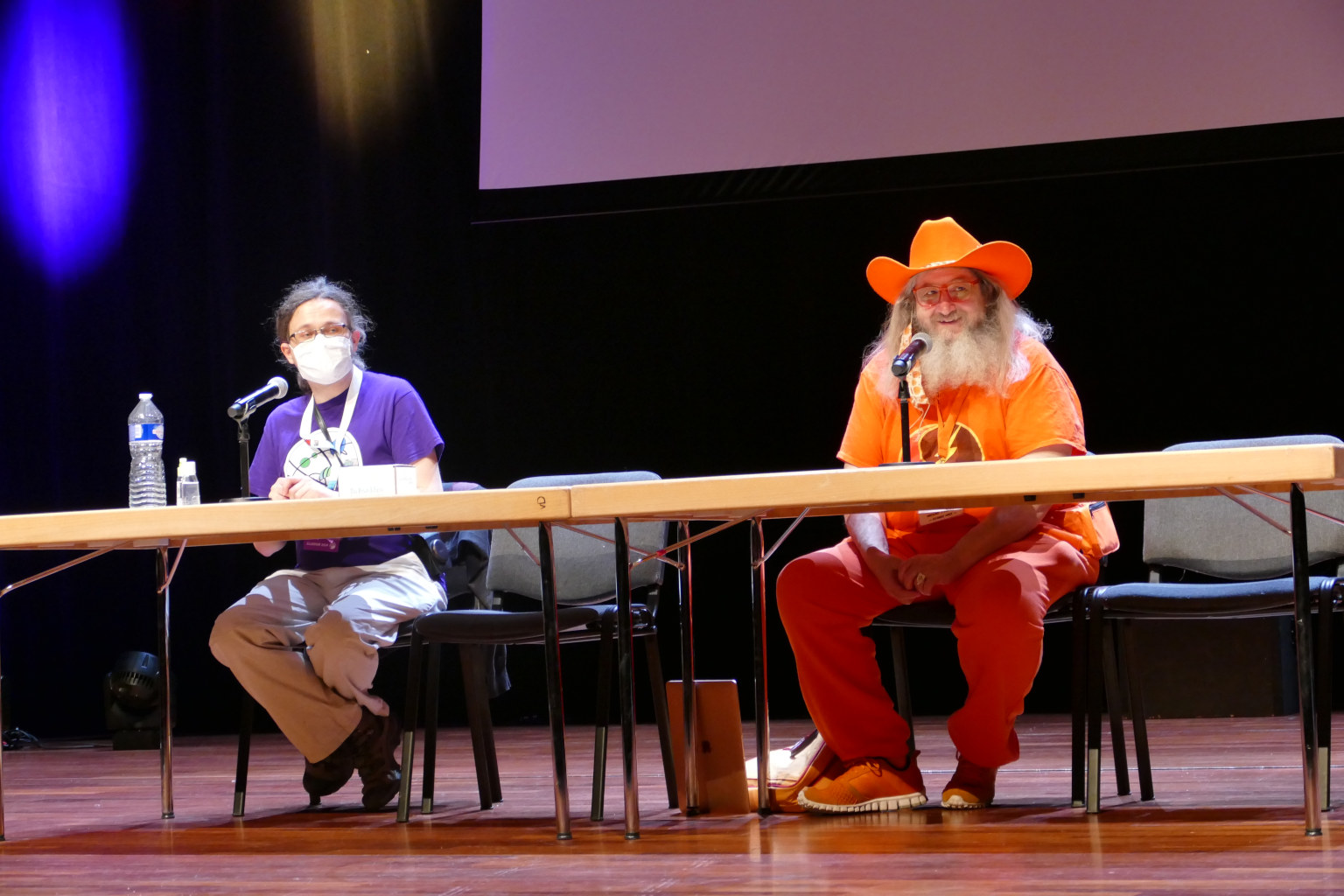 Two man sitting behind the tables. One wears mask. Second one is dressed fully in orange.