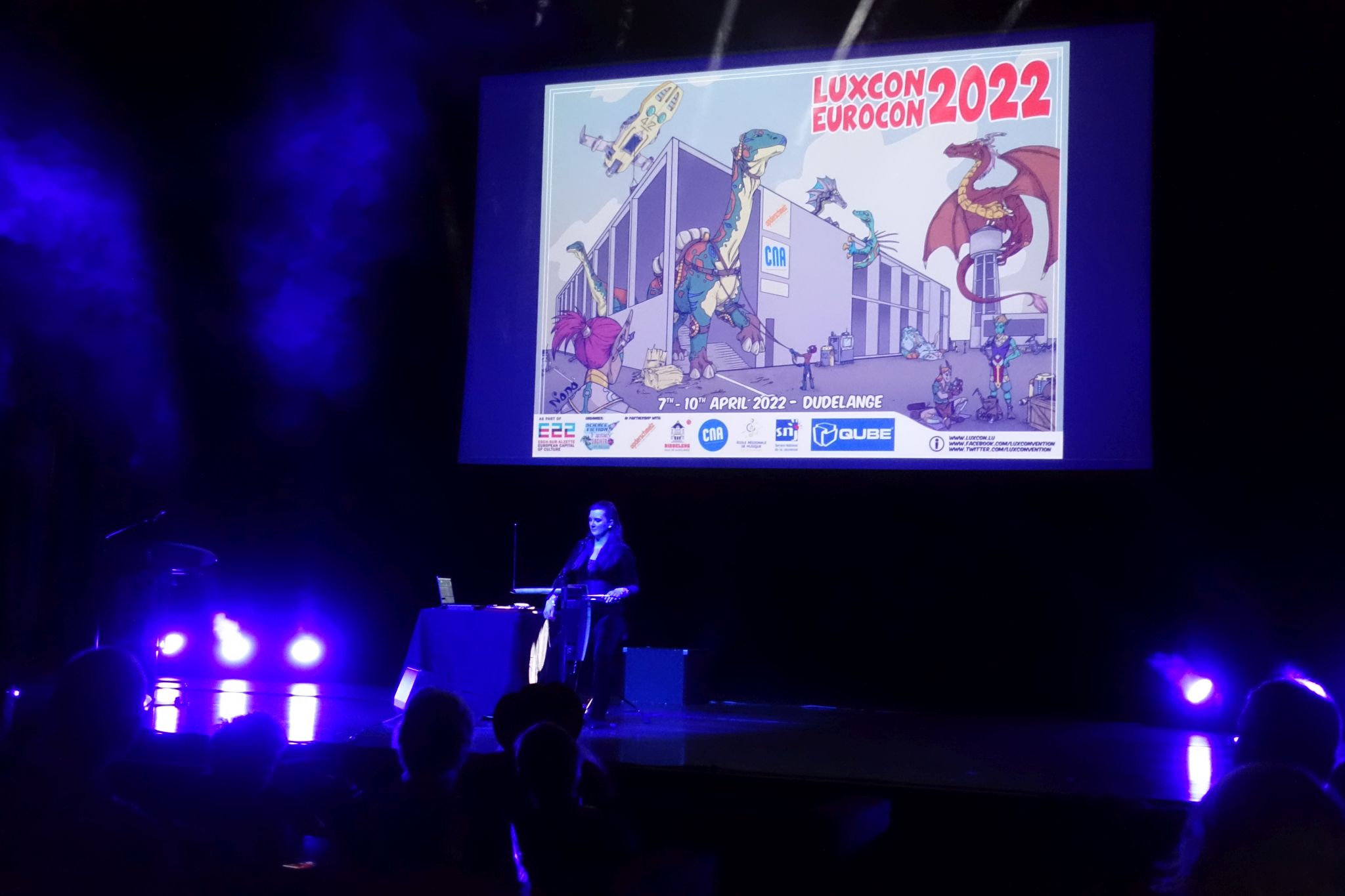 Women standing on stage in fron of the theremin. Behind her a there is a screen with convention graphic and name - Luxcon Eurocon 2022. The graphic presents dinosaurs and some fantastical creatures and vehicles around the convention venue. Whole picture has a blueish hue.