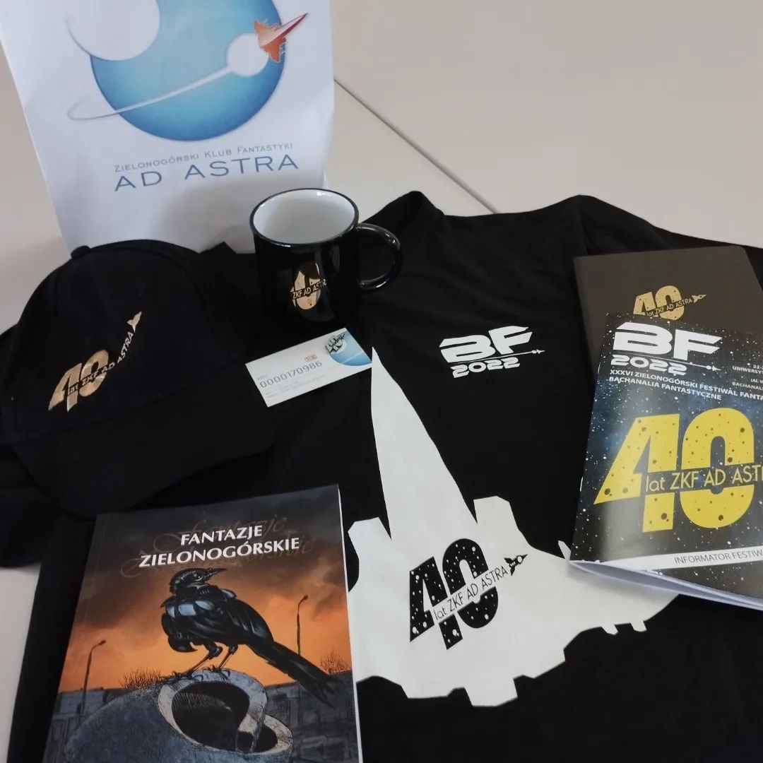 Picture depicts a t-shirt with a space fighter. There are two inscriptions 'BF 2022' and '40lat ZKF Ad Astra'. On the t-shirt there are other items (many with same description) baseball cap, a mug, a visiting card with a pin, programme booklet and black notebook. There is also a book with title 'Fantazje zielonogórskie'.