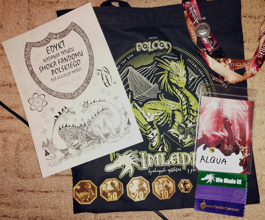 Picture shows multiple items from a convention. At the bottom there is bag with dragon and Polcon name. On top there is a lanyard and a badge with three ribbons, a set of wodden hexagonal coins and a diploma.