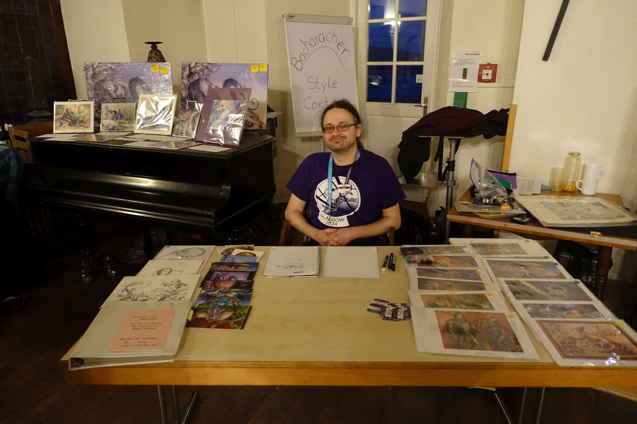 A man in purple t-shirt sits behind a table. There are art prints and posters on the table.