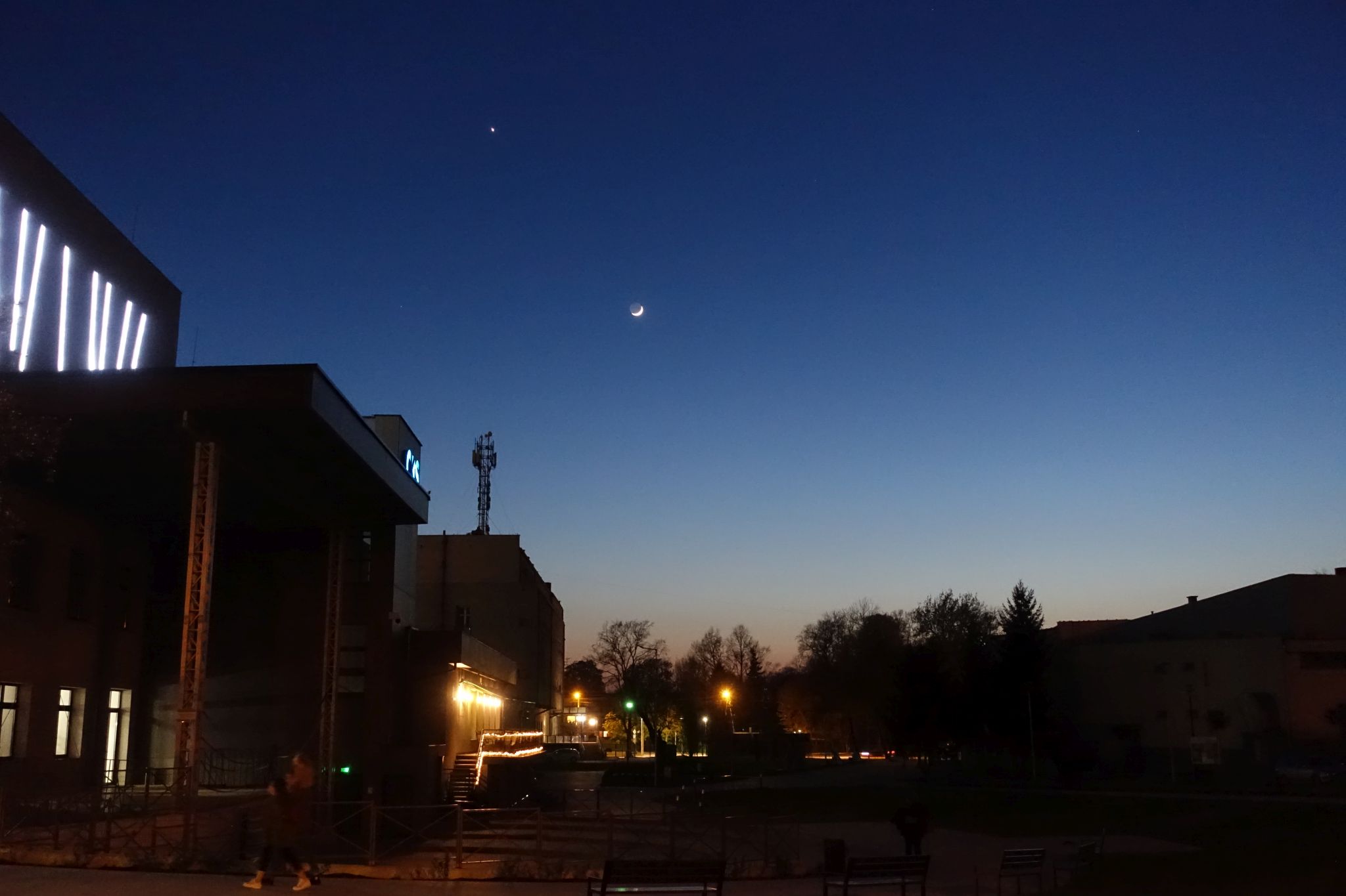 A sky just after sunset. Moon and Venus are clearly visible. Bottom and left part of the picture shows some buildings and trees.
