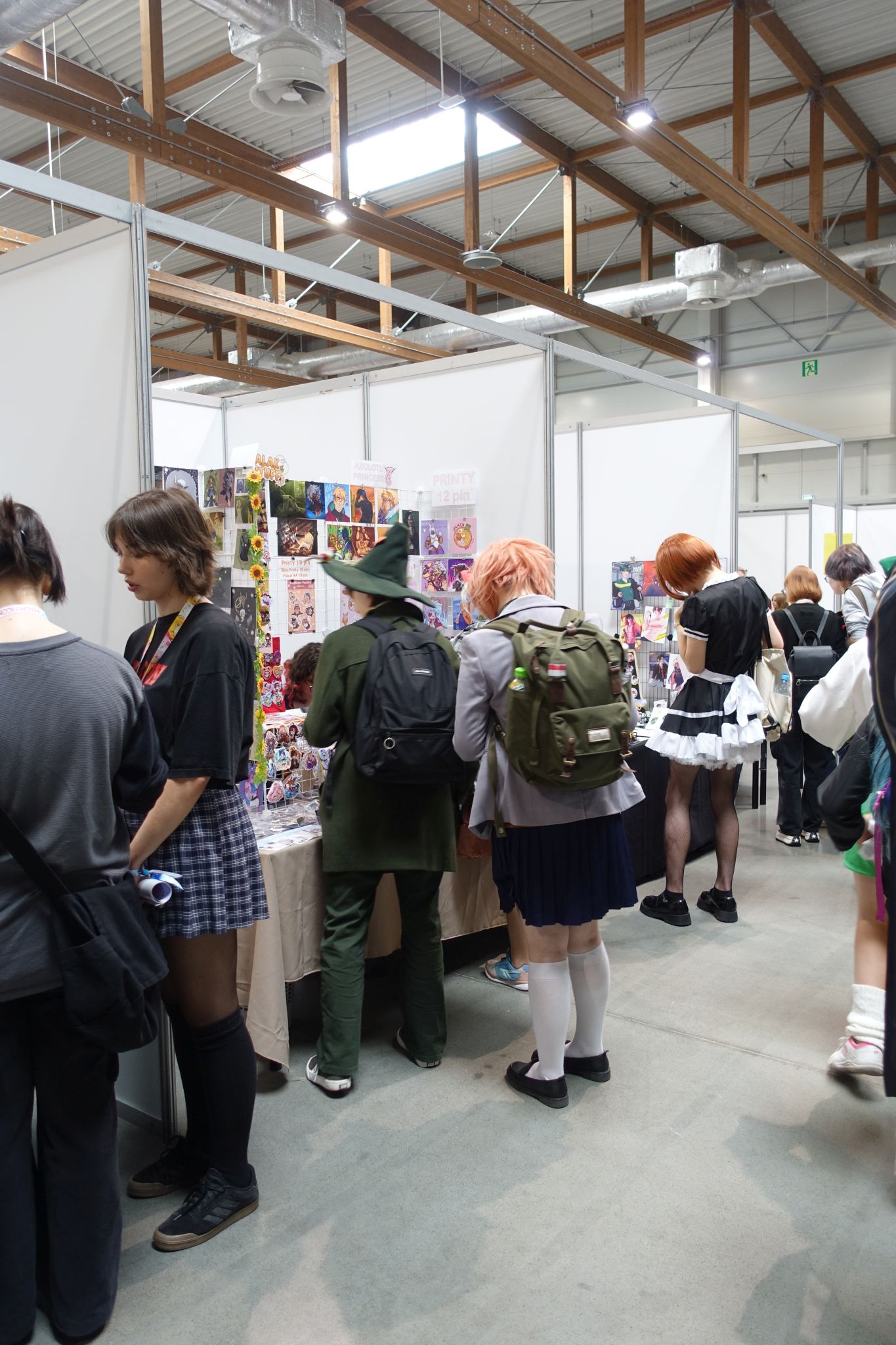 A few booths in the hall. In the center there is a person in the Snufkin costume. More people are standing in front of the booths.