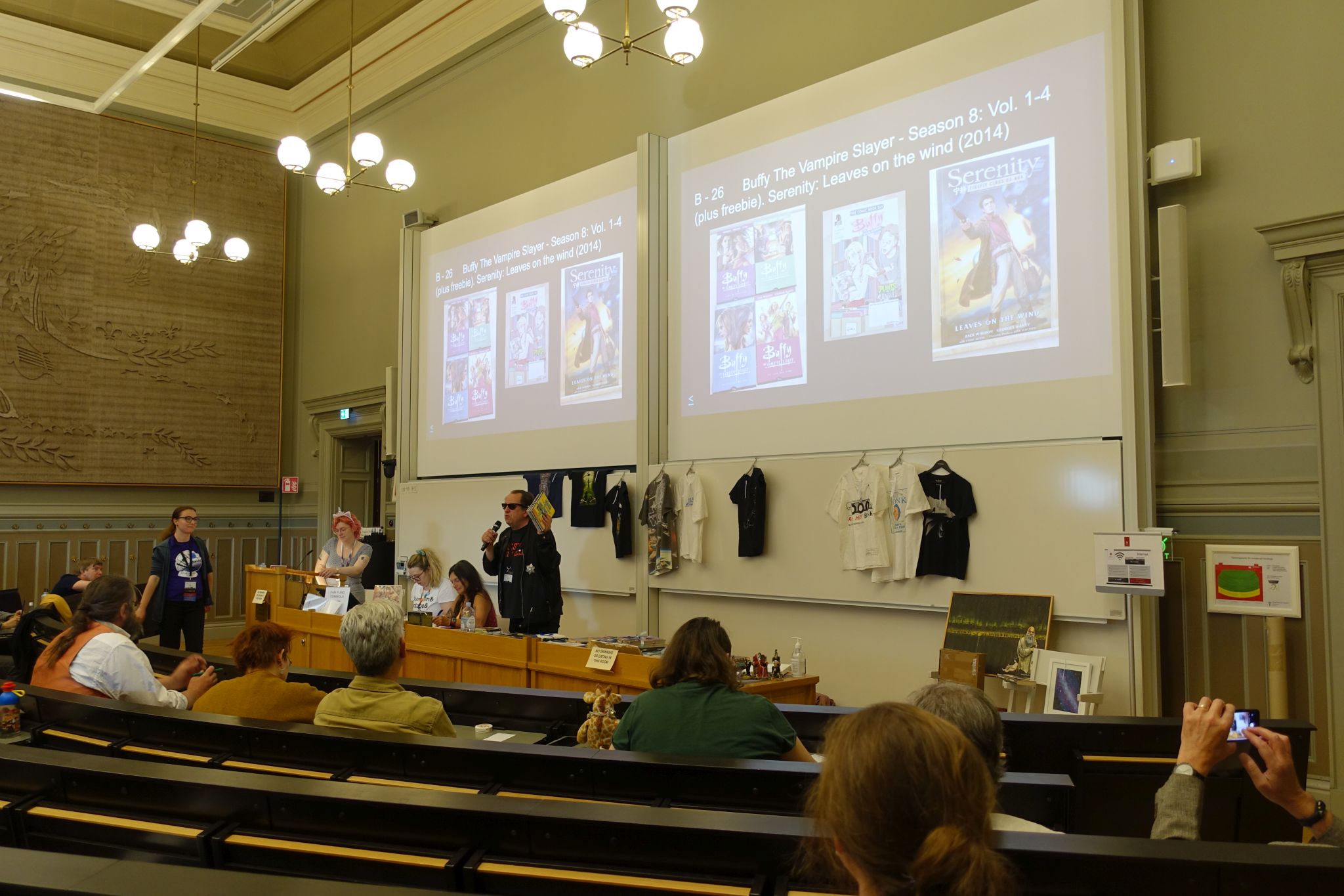 Picture taken in an auditorium style lecture hall. A few rows of seets are visible and some people are occupying chairs. On the middle of the picture there are few desks with people sitting and standing behind them. One person holds a microphone. behind them there are two screens with presentation showing some items on sale.