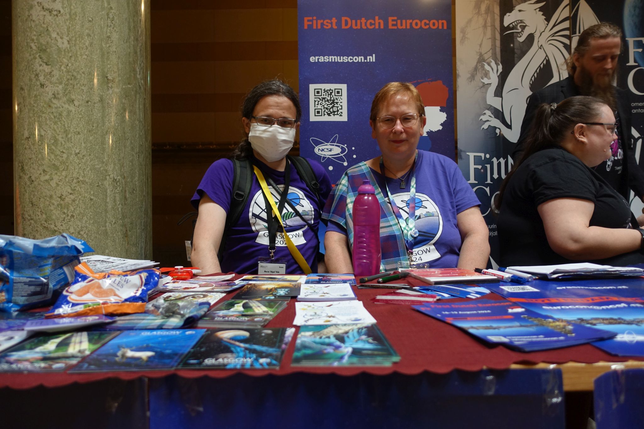 Two people posing for a picture behind a table with convention merchandise on it. Both are wearing purple t-shirts with Glasgow 2024 logo. Next to them there are two more people.