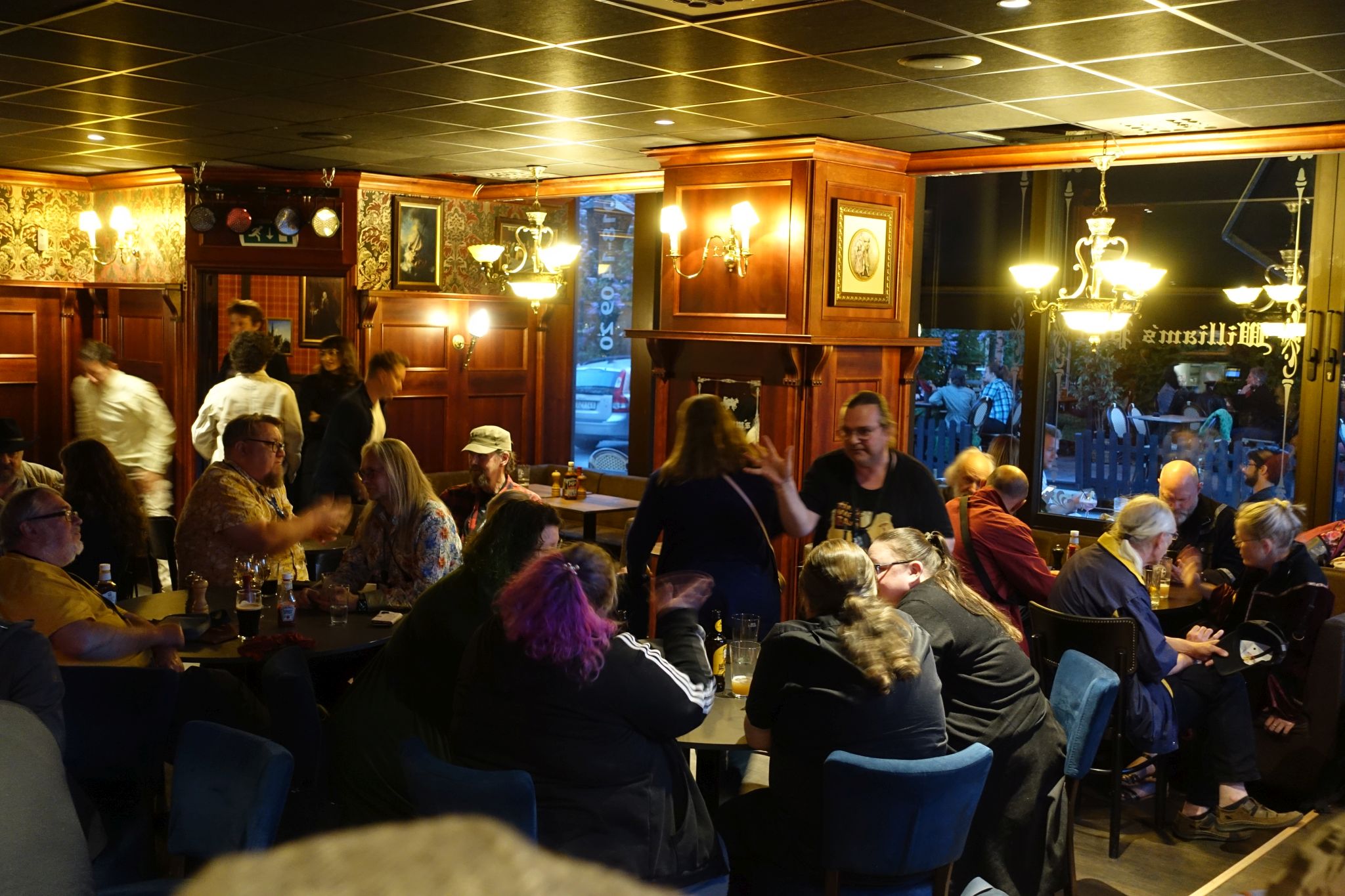Interior of the pub. There are many people sitting and standing. The walls are wooden and there are big windows. Outside there are more tables and people sit around them.