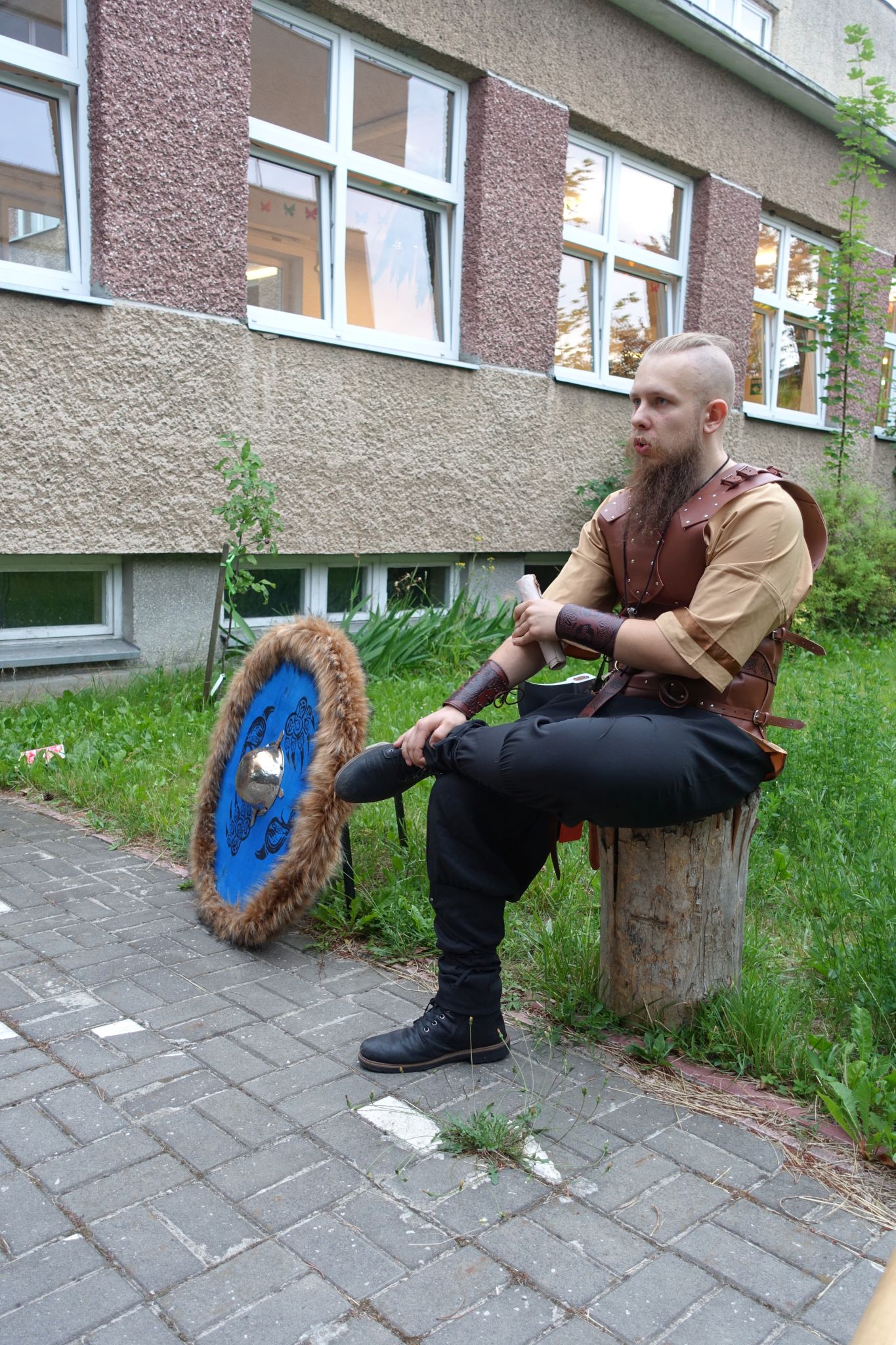 Man dressed in a wooden armor sits on a stump. Next to him there is a round, blue shield.