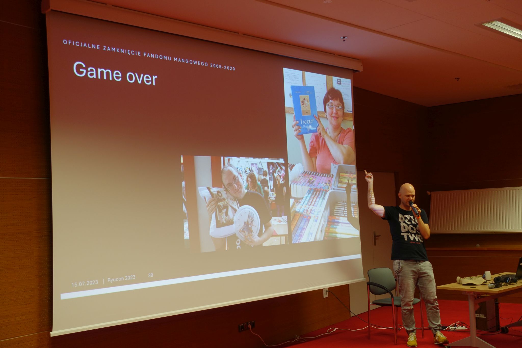 A man speaking to the microphone and pointing his finger to the screen behind him. The screen shows pictures of two people and the text 'Game over'.