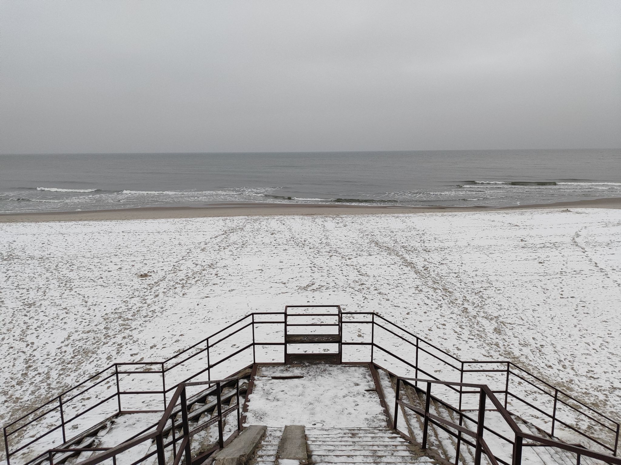 A view of sea and the beach as visible from the top of the stairs. Stairs and part of the beach are covered in snow.