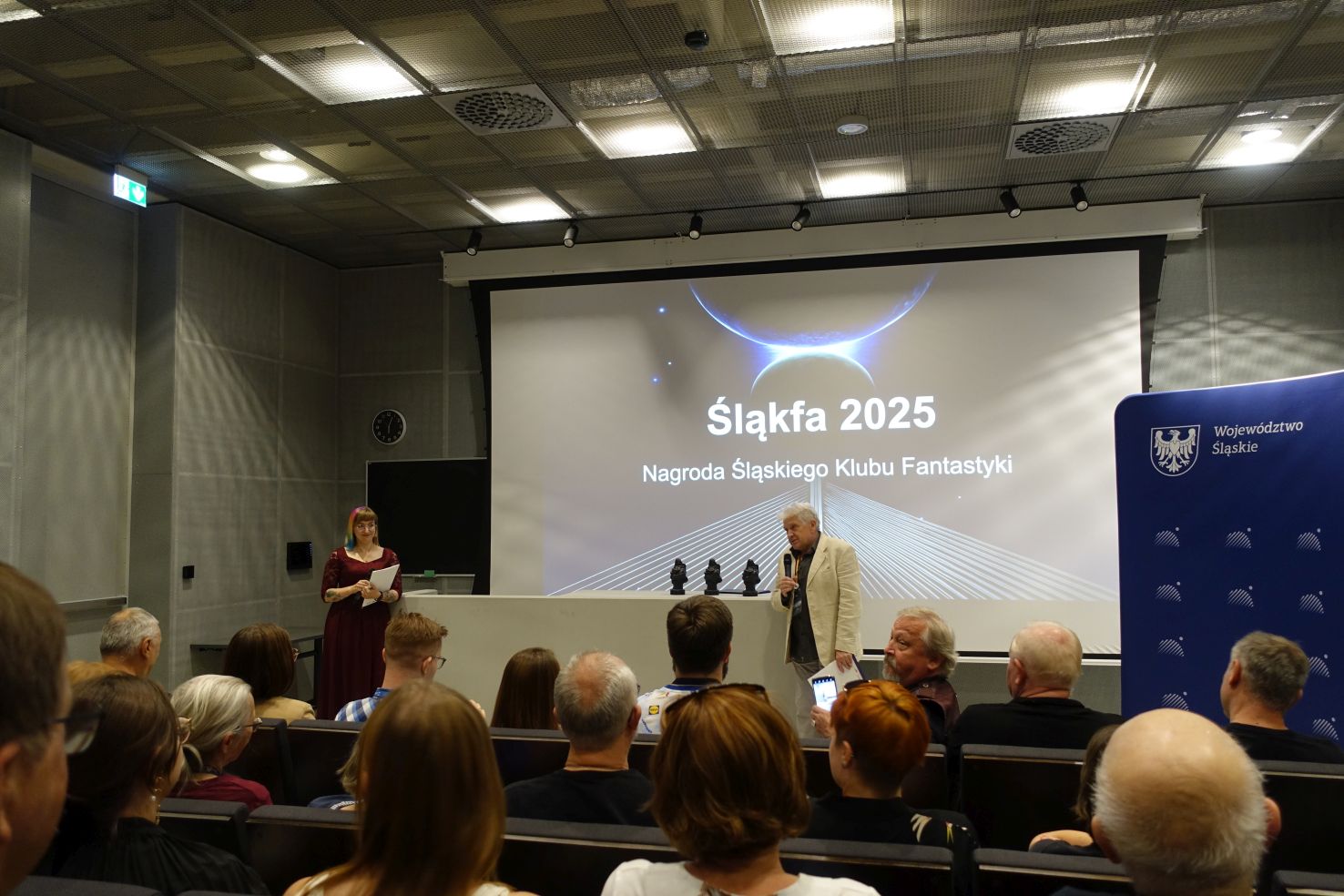 A woman and a men in official clothes stand in front of the audience. Behind them there is a screen with a picture and a text. The text reads 'Śląkfa 2025' and below 'Nagroda Śląskiego Klubu Fantastyki'.